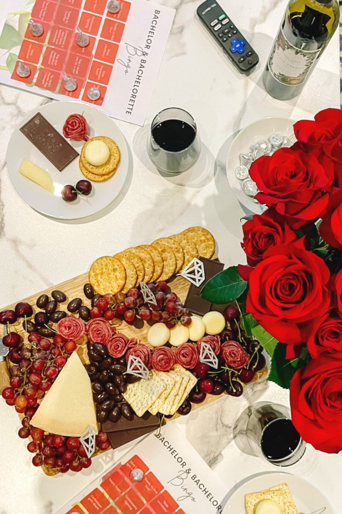 Overhead image of engagement charcuterie board on coffee table with bingo card, wine, and flowers